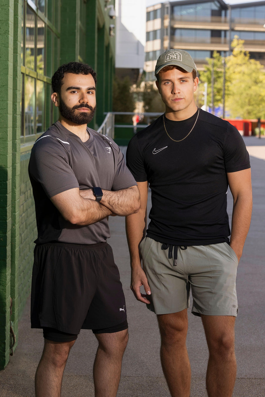 Two men standing outdoors, one wearing a gray shirt and black shorts, the other in a black shirt and gray shorts, with a building and trees in the background.