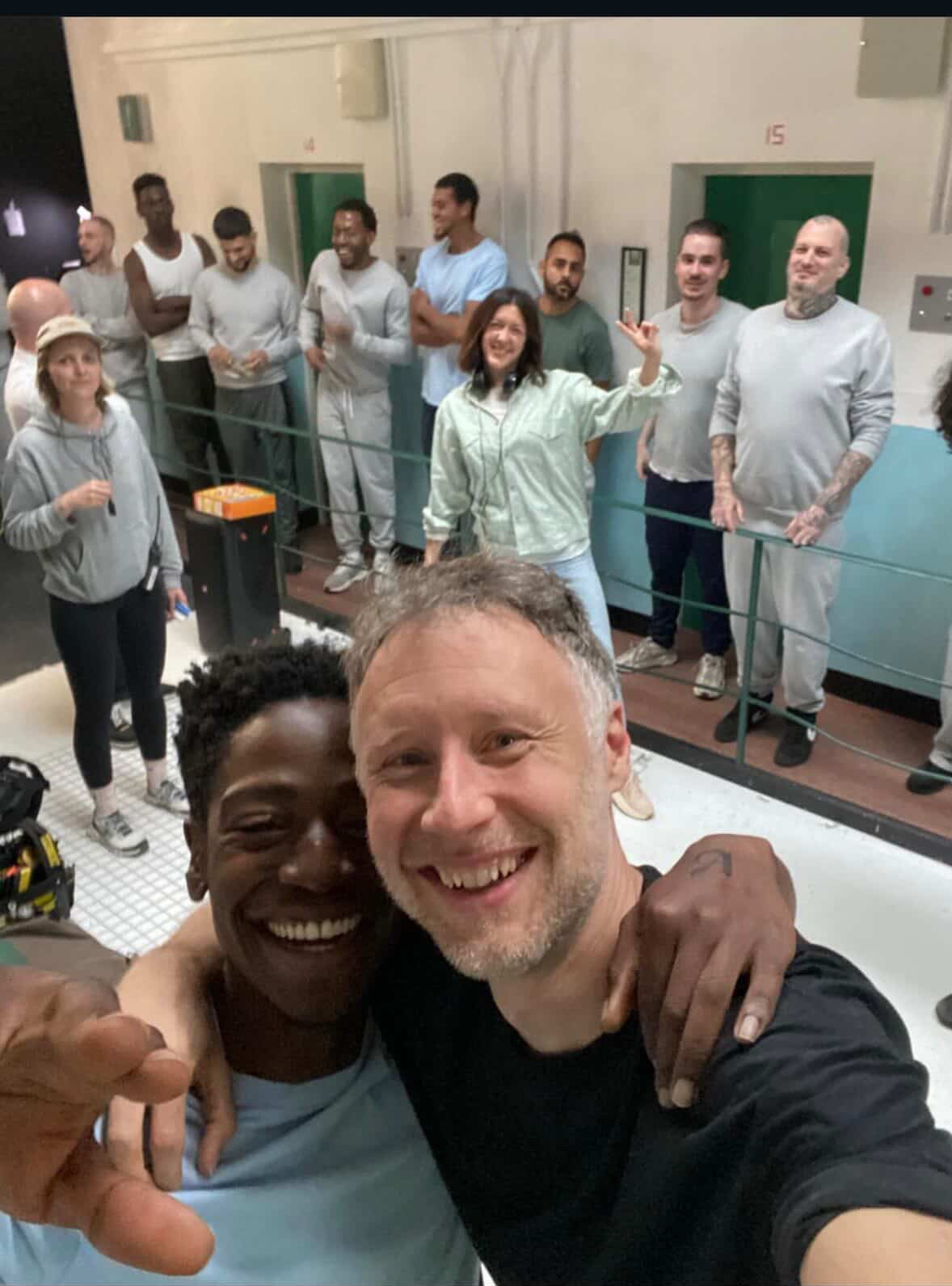 Two men taking a selfie with a group of people in a prison setting. Behind the scenes on set for the filming of Wasteman