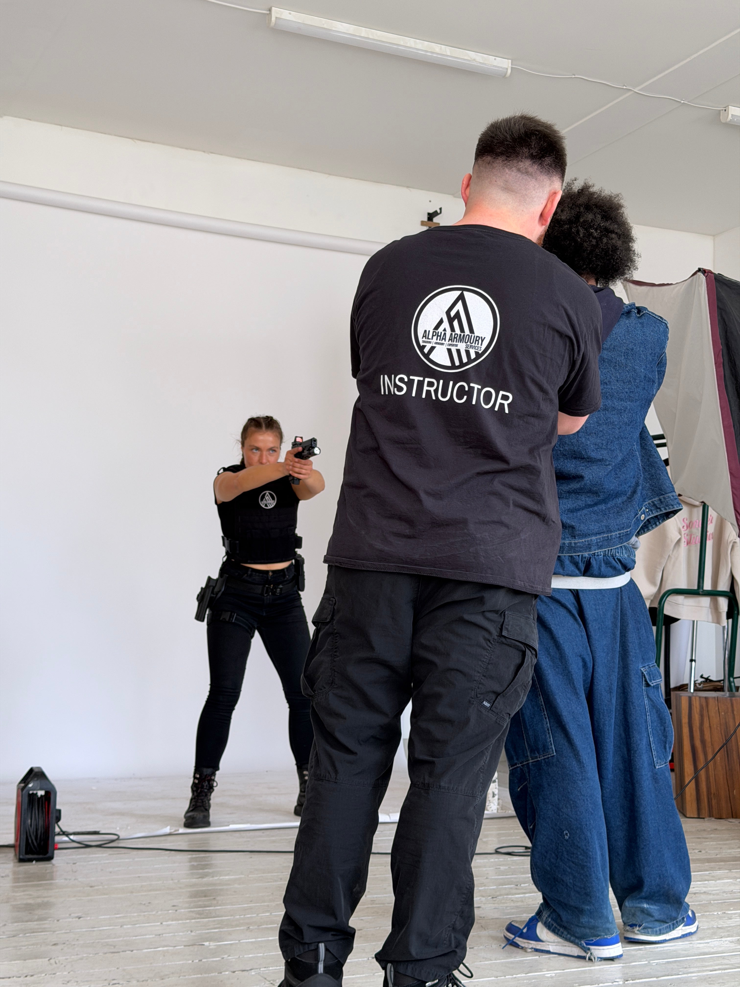 Three people behind the scenes of our Sonder Character Profile shoot in a room with one wearing an 'Instructor' jacket, one is a photographer taking a picture of the third holding a gun with a white backdrop