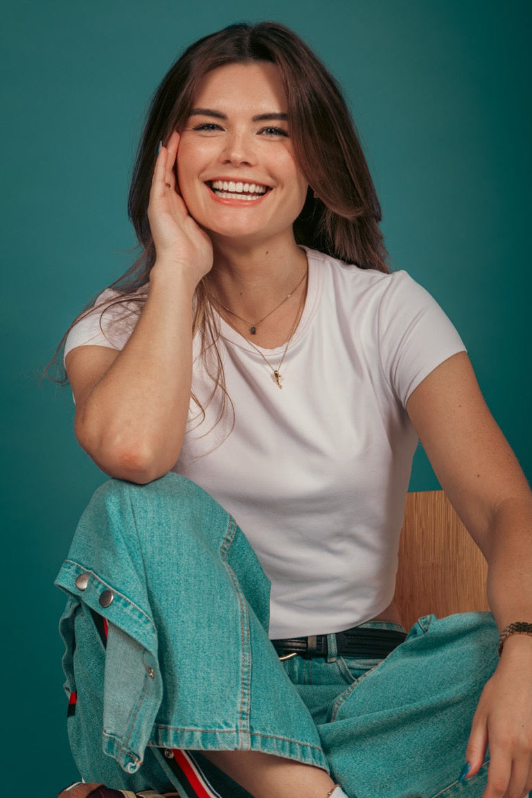A smiling woman sits cross-legged on a wooden chair against a teal background, wearing a white t-shirt, light blue denim jeans, brown sneakers, and white socks.