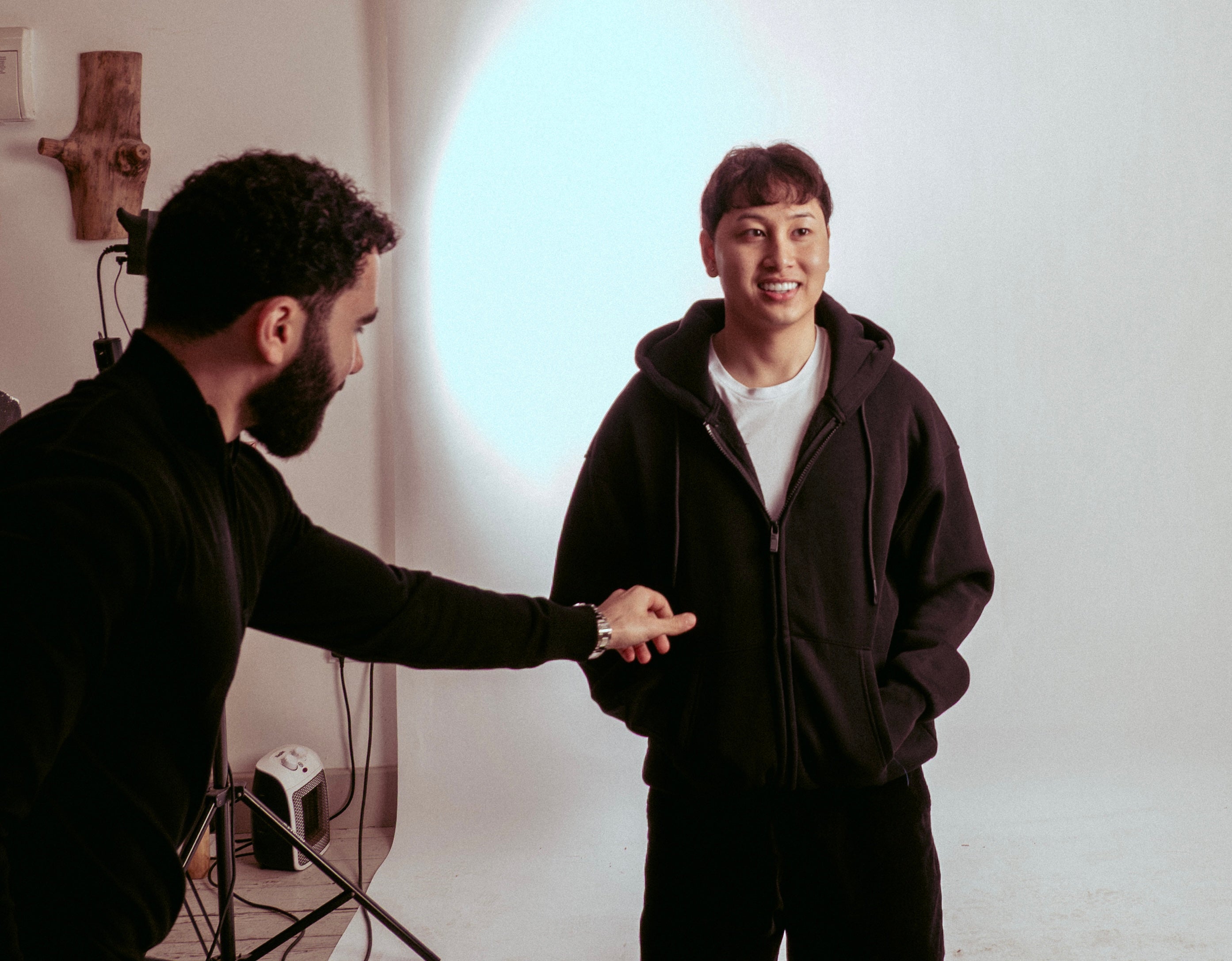 Two men in a studio setting whilst one fixes the positioning of the others hoodie