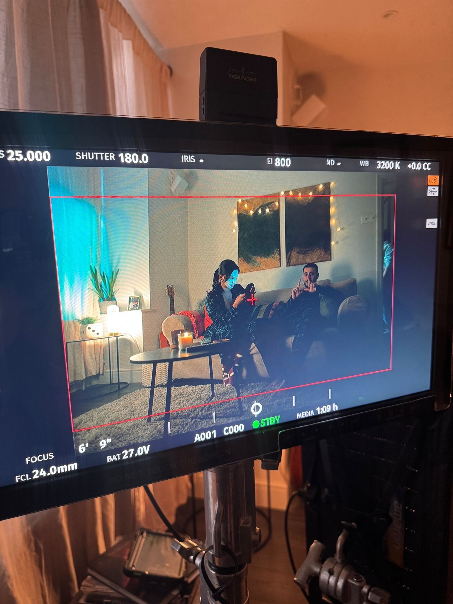 Camera monitor displaying a scene of two people in a living room with filming equipment around.