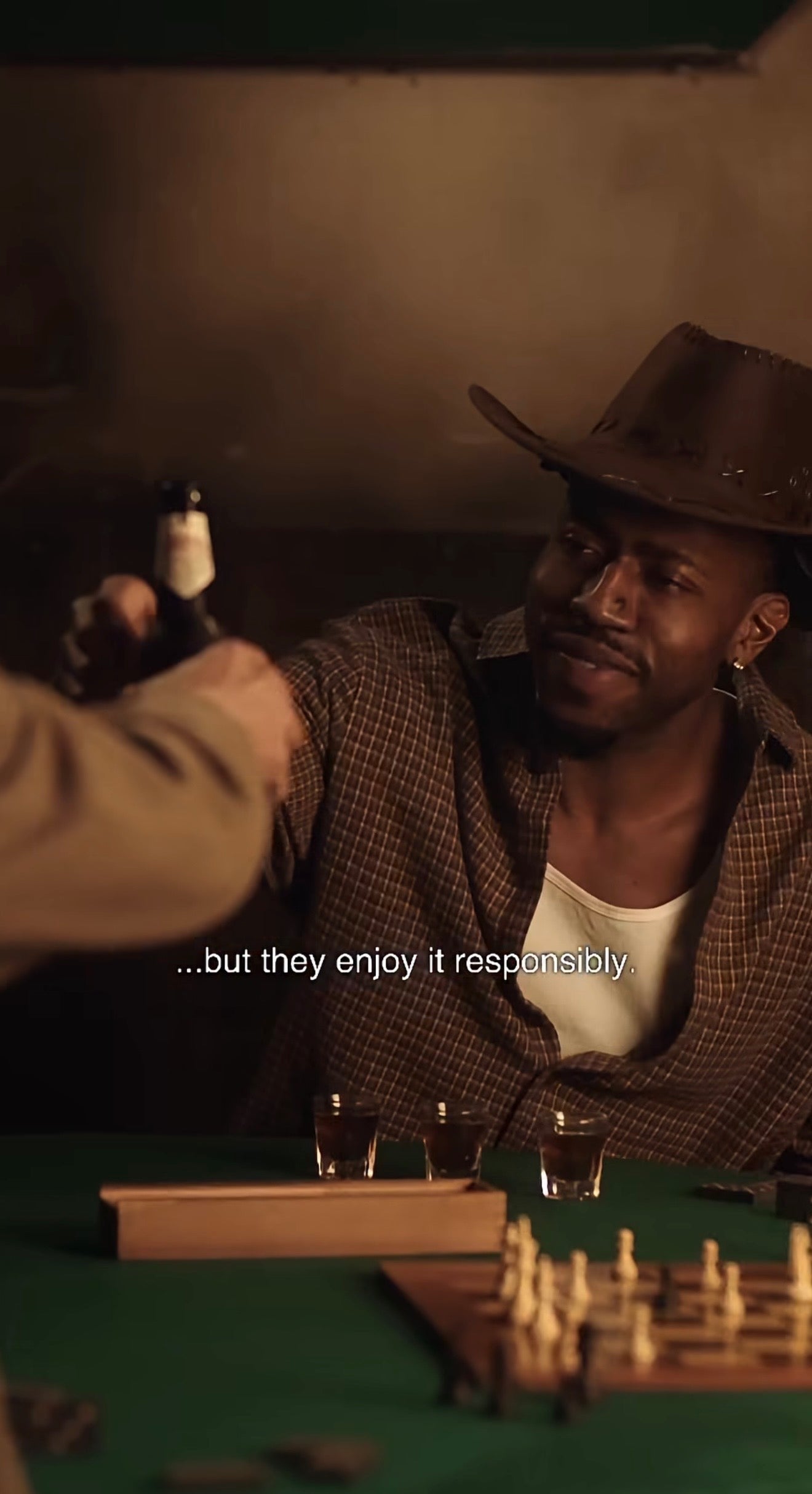 Remi is the Man in a cowboy hat sitting at a table giving cheers to another man with drinks while a chess game and pieces are on the table, with text overlay. Taken from a shoot Remi was featured in. 