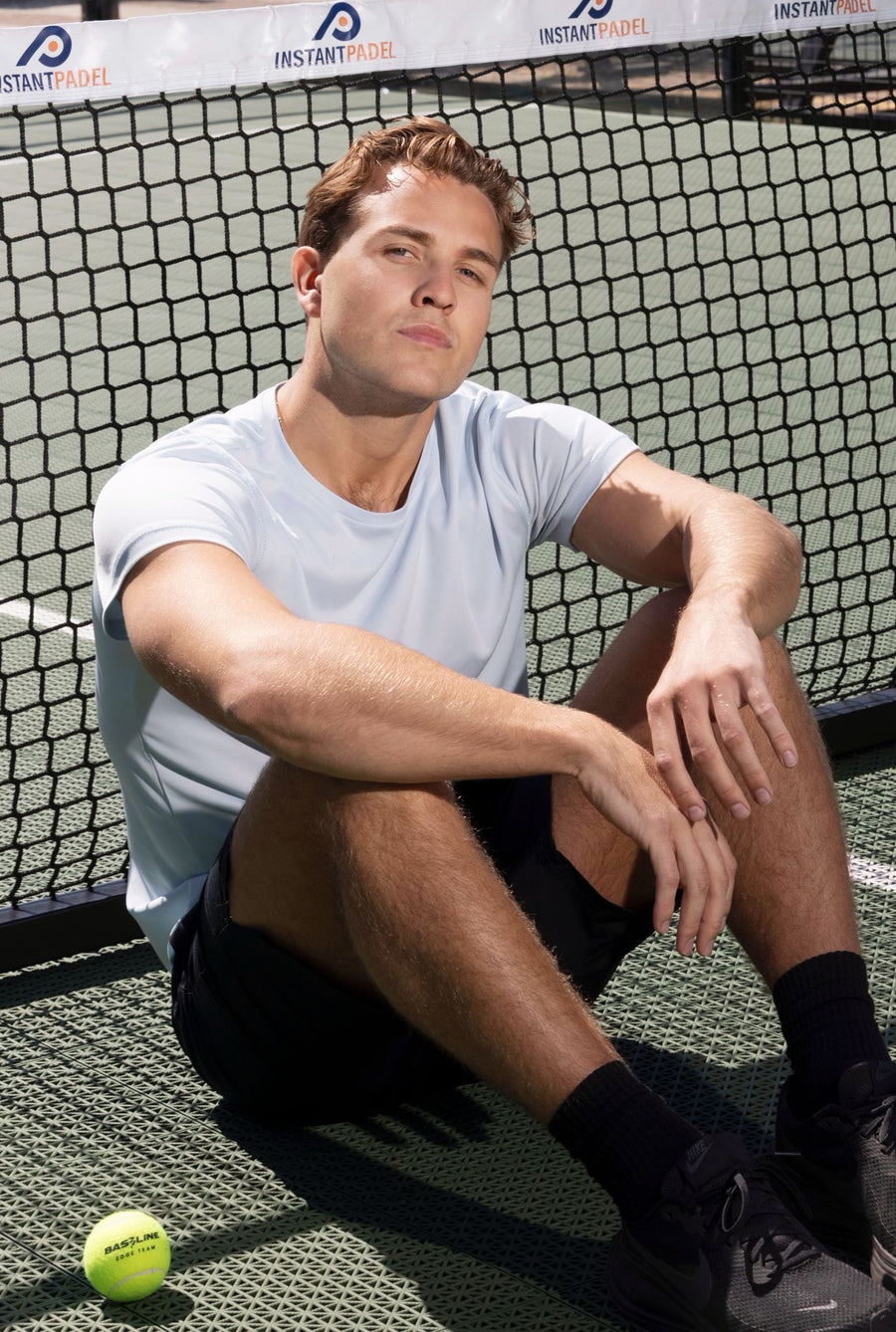 Man sitting on a tennis court with a tennis ball next to him, wearing a blue t-shirt and black shorts.