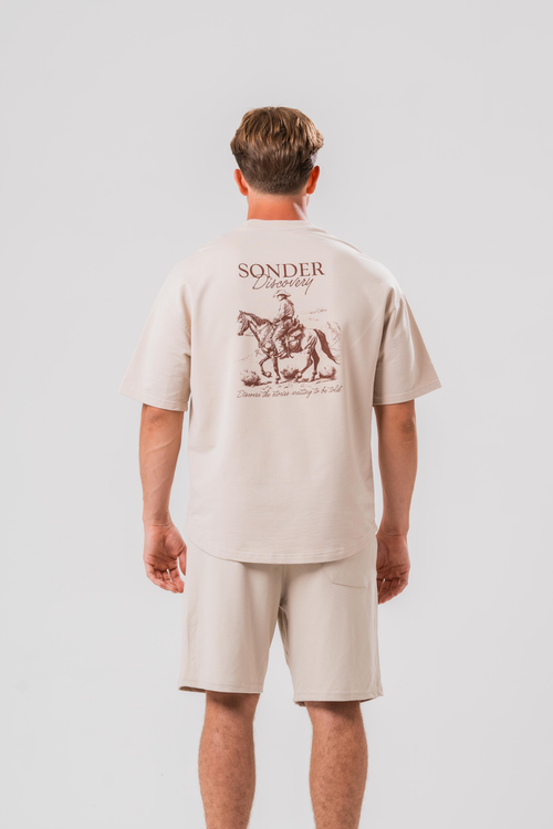 Back of a Male model wearing the Sonder Beige cowboy tee and beige shorts, in front of a light grey background