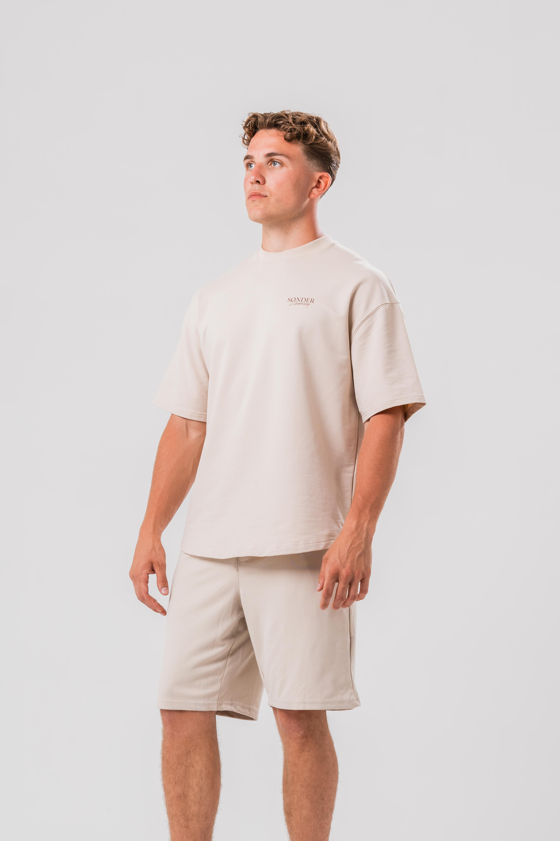 Side view of Male model wearing the Sonder Beige cowboy tee and beige shorts, in front of a light grey background