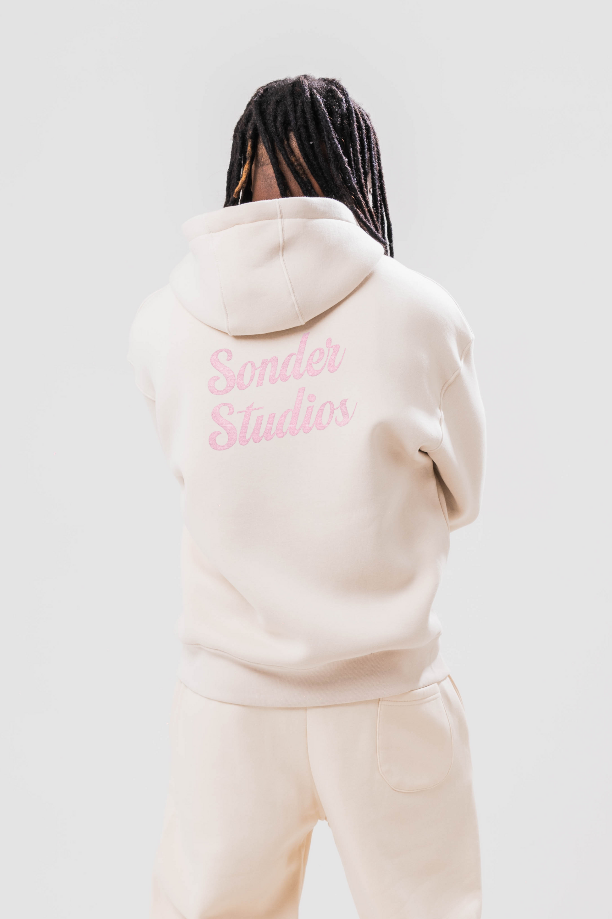 Back view of Male model wearing the Sonder Beige Oversized zip hoodie with the pink Sonder Studios design paired with the oversized joggers. Standing in front of a light grey background.