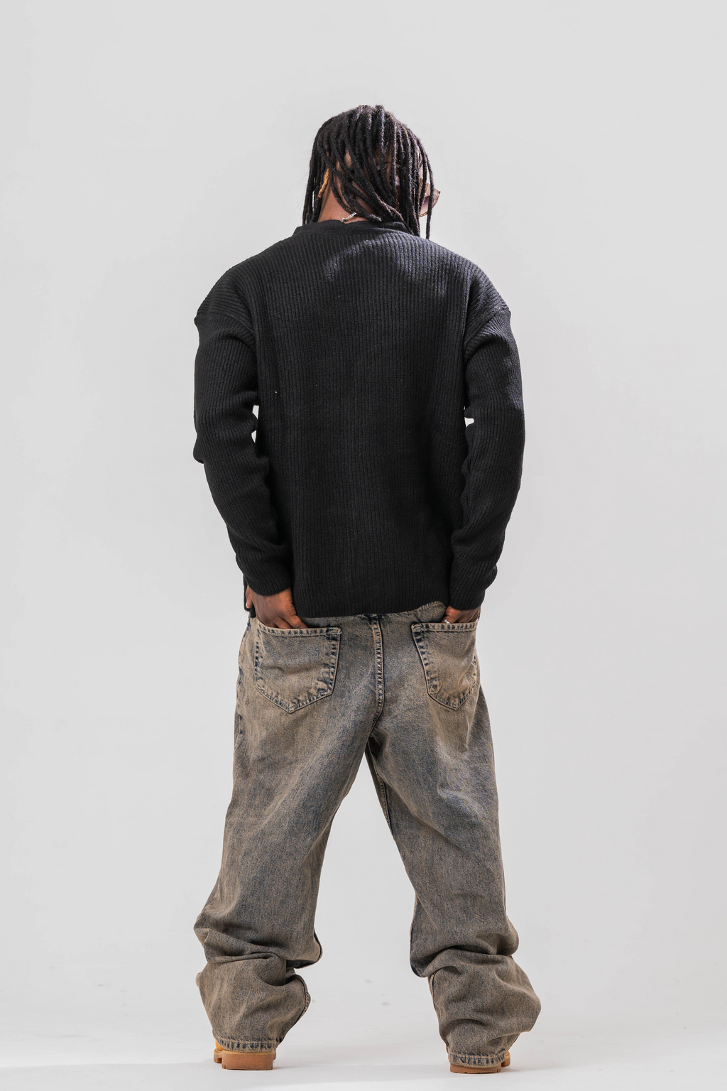 Back view of Male model wearing the Sonder Black Knit Cardigan with baggy jeans and sun glasses. In front of a light grey background.