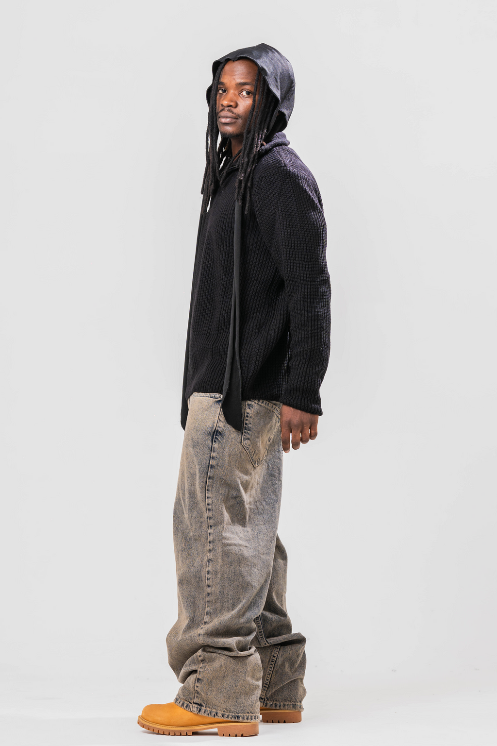 Side view of Male model wearing the Sonder Black Knit Quarter Zip with baggy jeans and a durag. In Front of a light grey background.