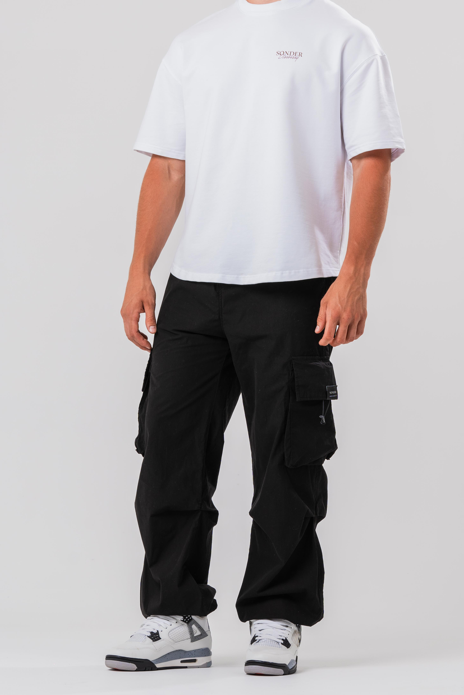 Side view of Male model wearing the Sonder Black Oversized Cargo Pants and Sonder White Oversized Graphic Cowboy T-Shirt. In front of light grey background.