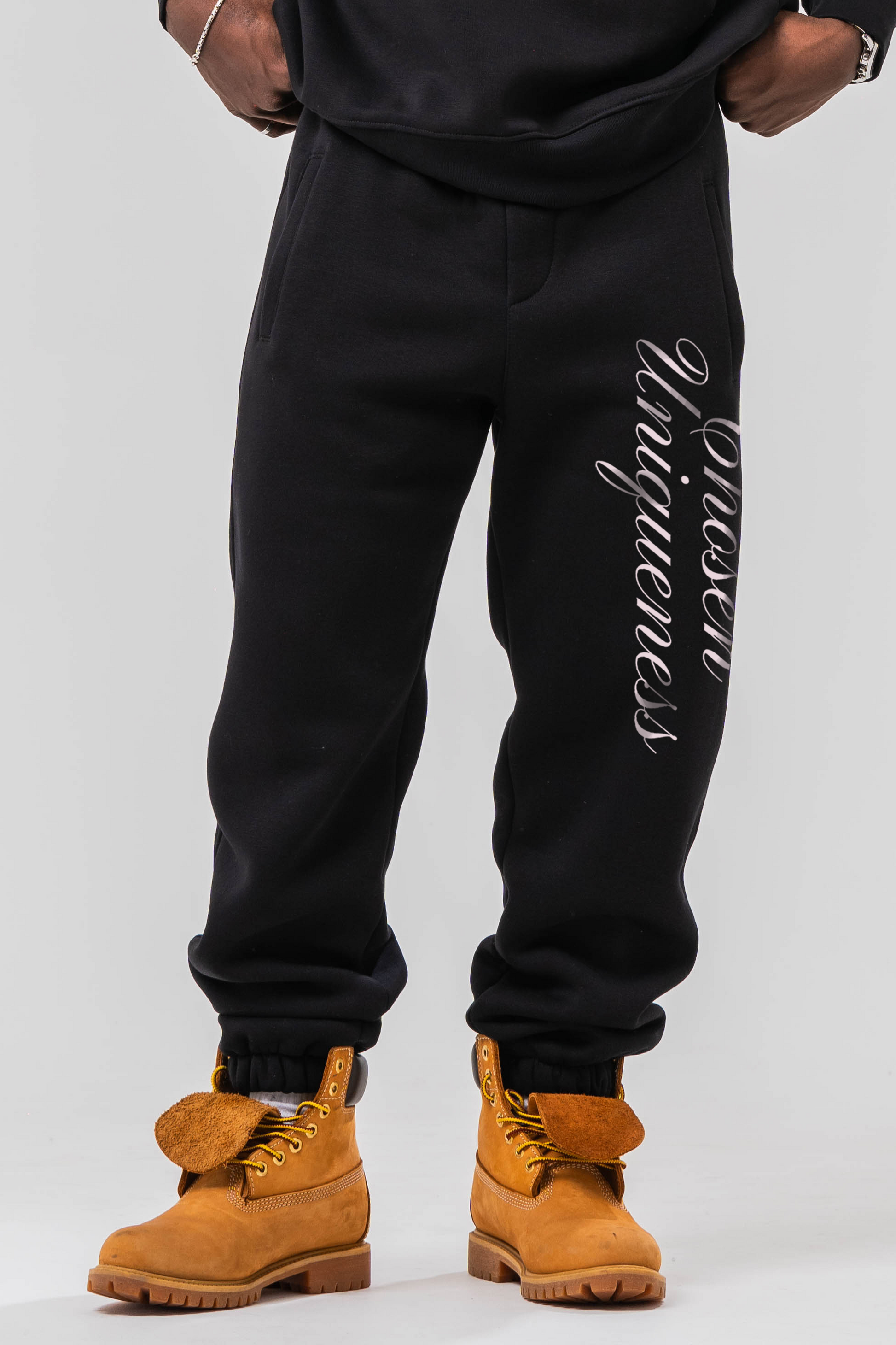 Male model wearing the Sonder Black Oversized sweatshirt with the oversized joggers with the Chosen Uniqueness print down the leg. Standing in front of a light grey background.