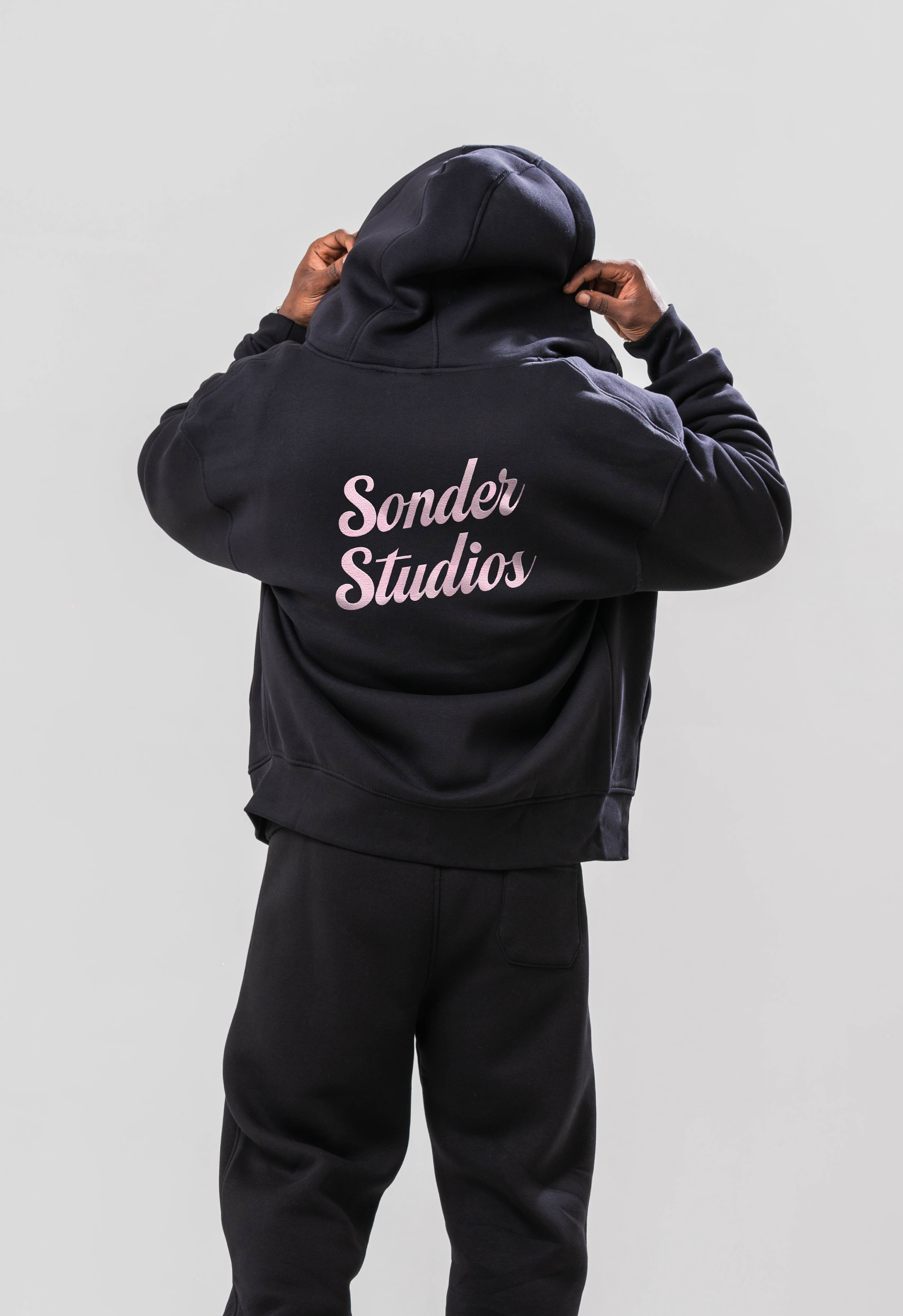 Male model wearing the Sonder Black Oversized zip hoodie with the Sonder Studios Pink logo on the back paired with the oversized joggers and black tee. Standing in front of a light grey background.