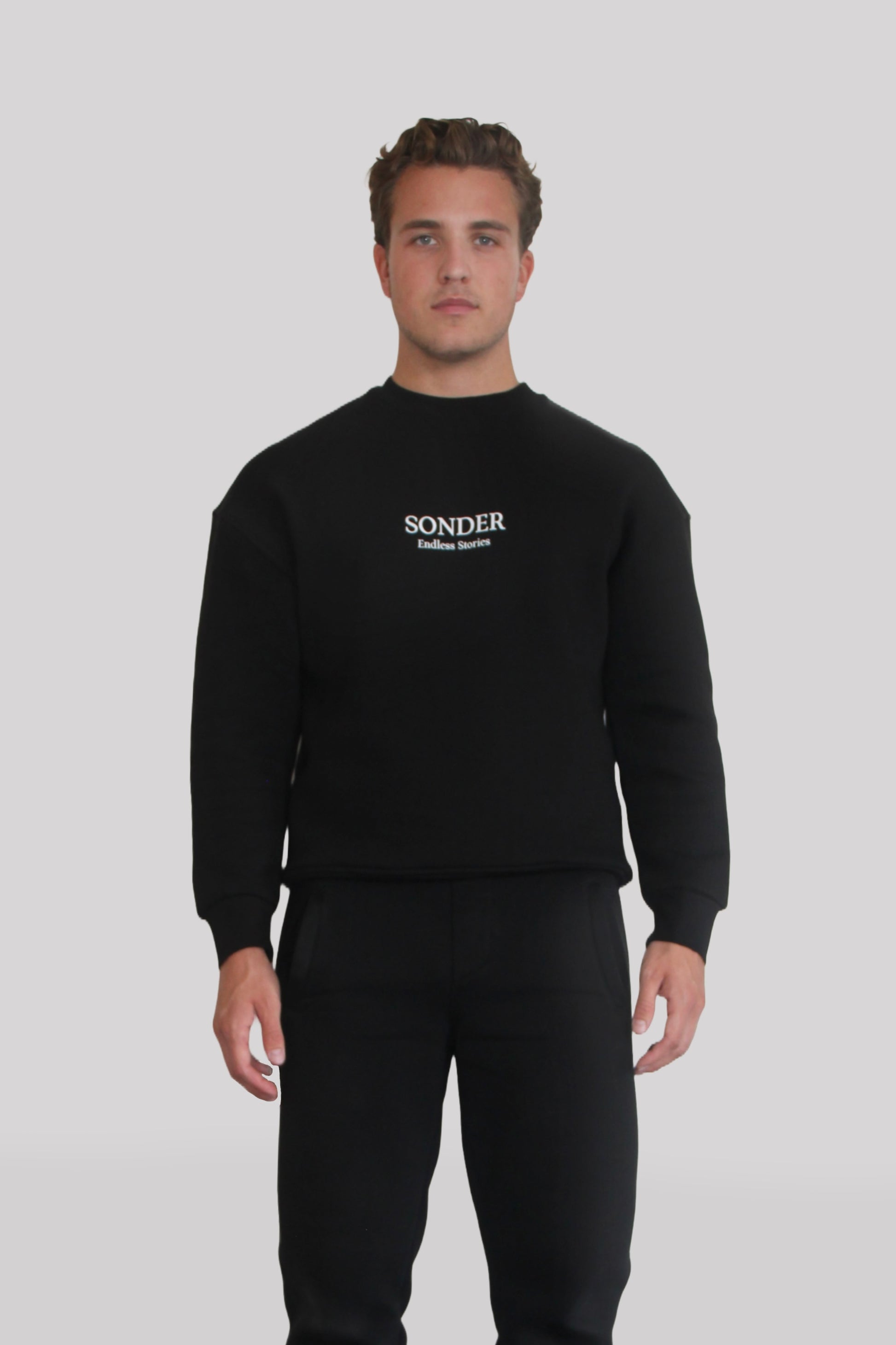 Male model wearing the Sonder Black Signature Series Sweatshirt with SONDER branding paired with the Black Signature Series Sweatpants. In front of a light grey background.
