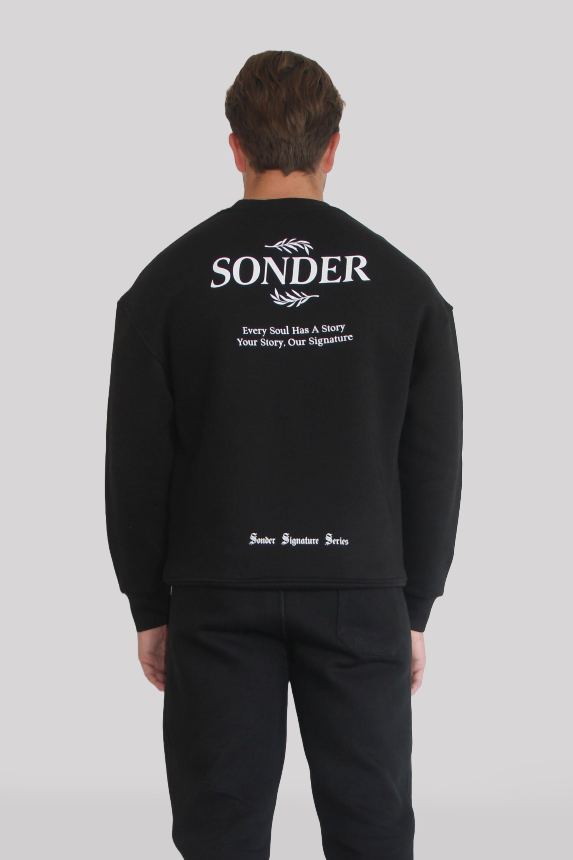 Back view of Male model wearing the Sonder Black Signature Series Sweatshirt with SONDER branding paired with the Black Signature Series Sweatpants. In front of a light grey background.