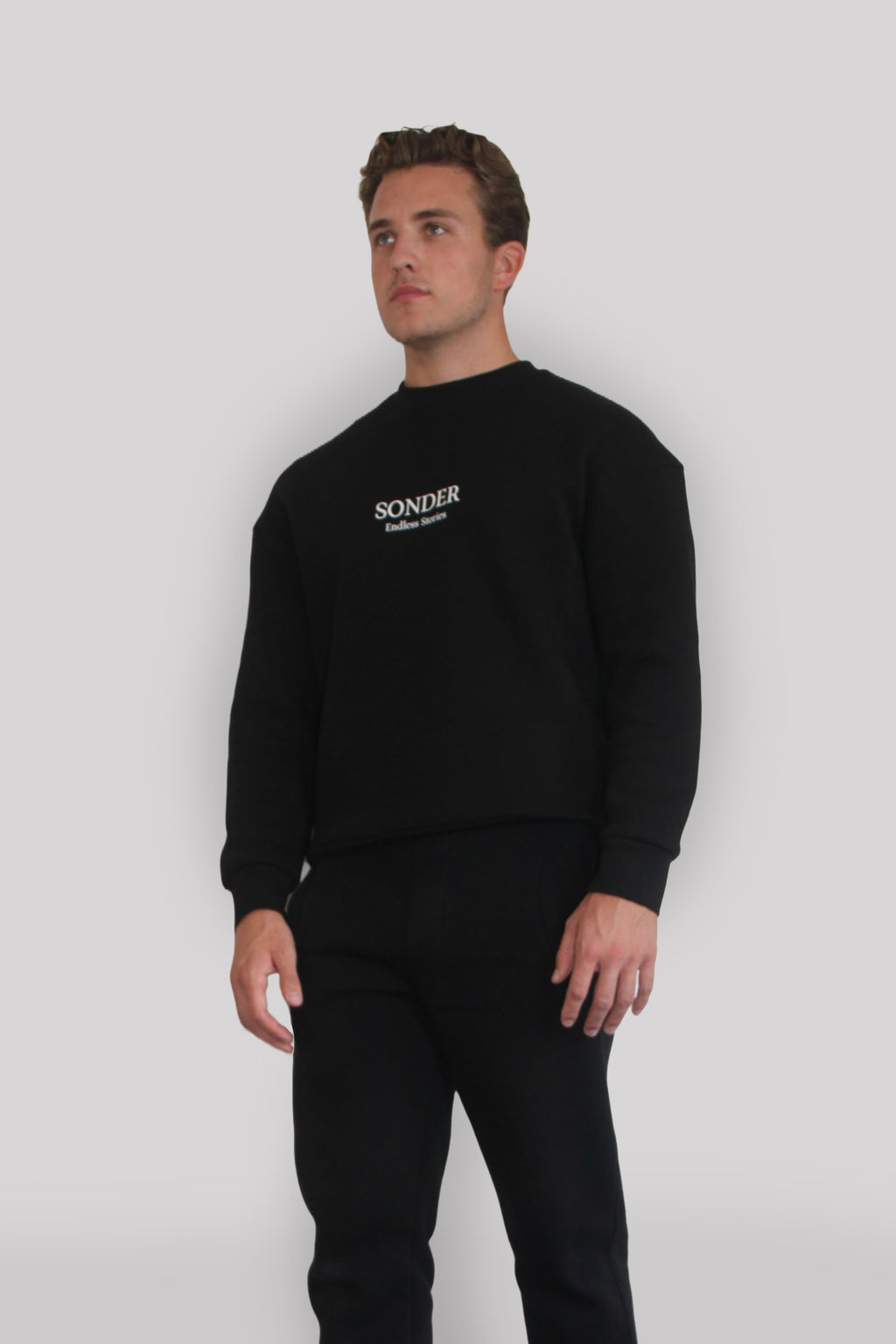Side view of Male model wearing the Sonder Black Signature Series Sweatshirt with SONDER branding paired with the Black Signature Series Sweatpants. In front of a light grey background.