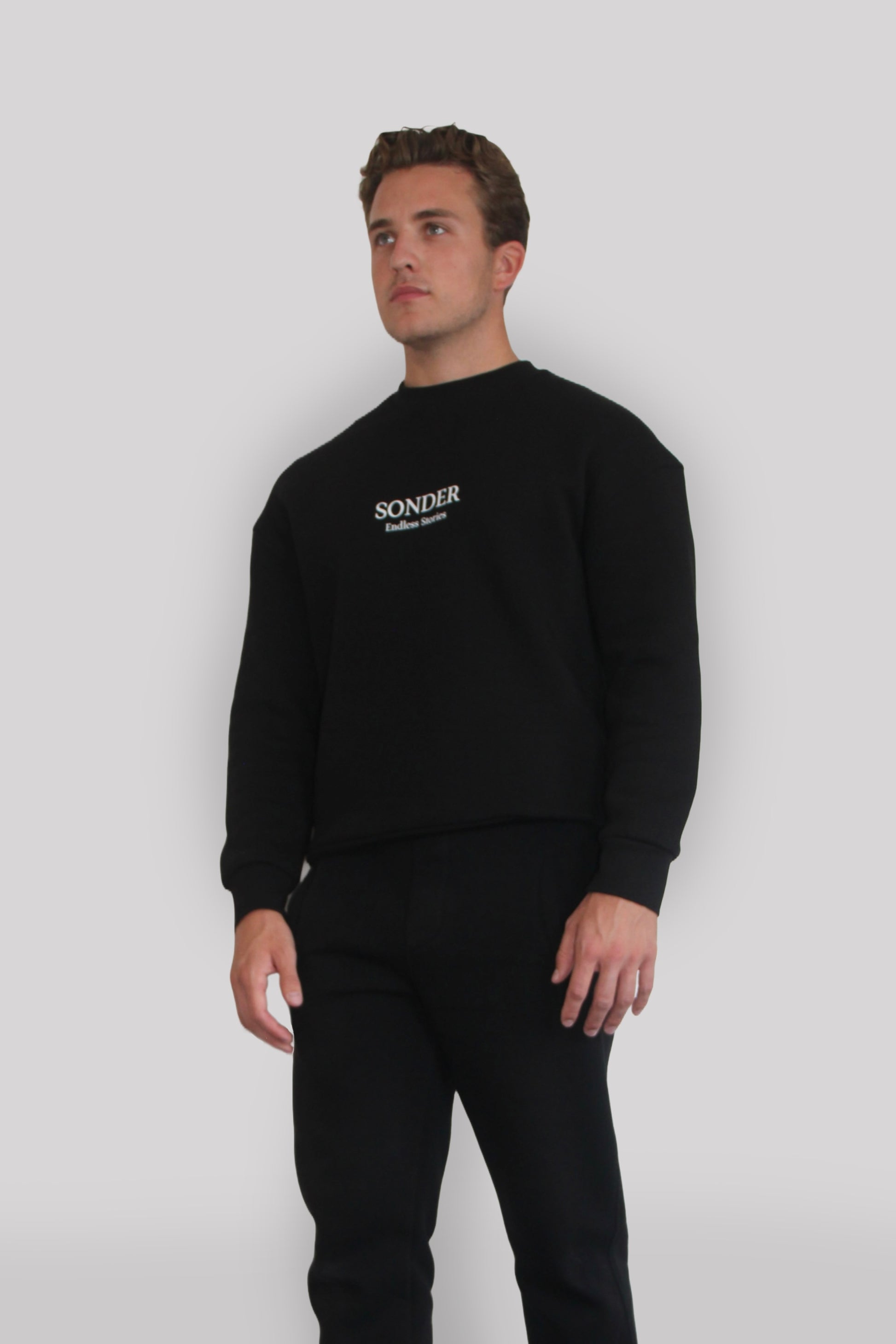 Side view of Male model wearing the Sonder Black Signature Series Sweatshirt with SONDER branding paired with the Black Signature Series Sweatpants. In front of a light grey background.