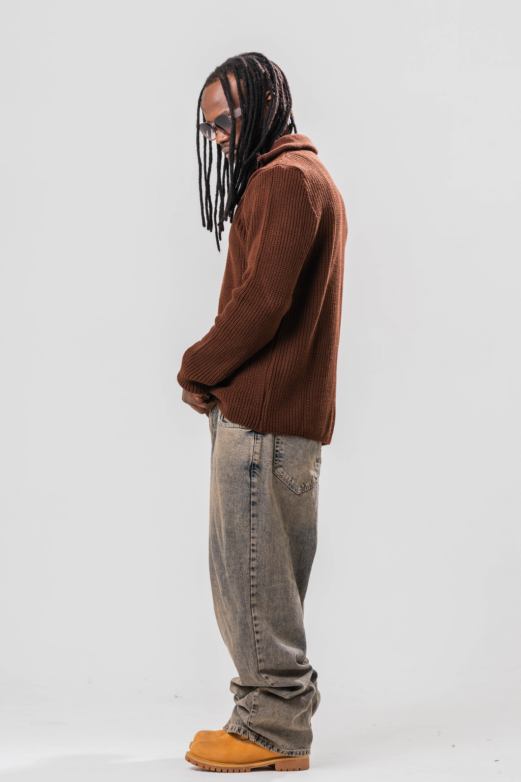 Side view of Male model wearing the Sonder Brown Knit Quarter Zip with baggy jeans and sun glasses. In front of a light grey background.