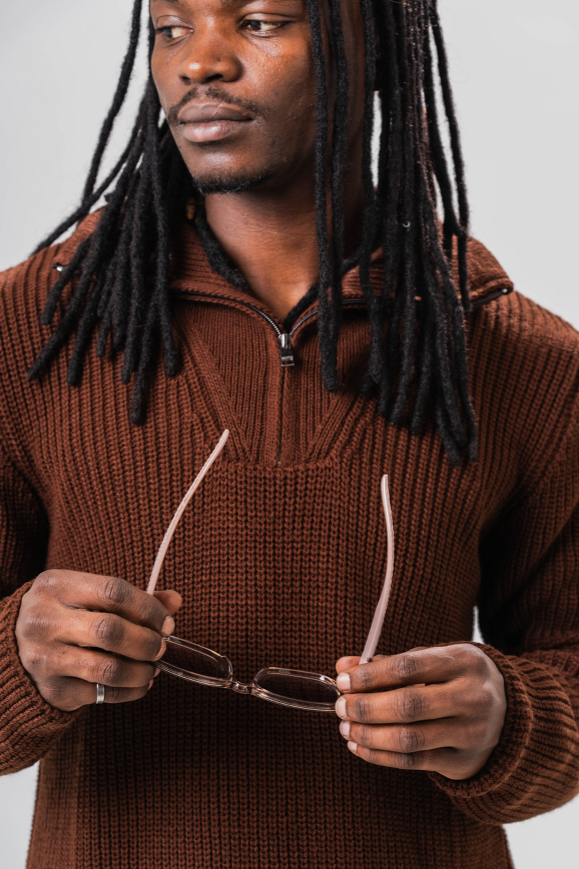 Close up of Male model wearing the Sonder Brown Knit Quarter Zip holding a pair of sunglasses. In front of a light grey background.