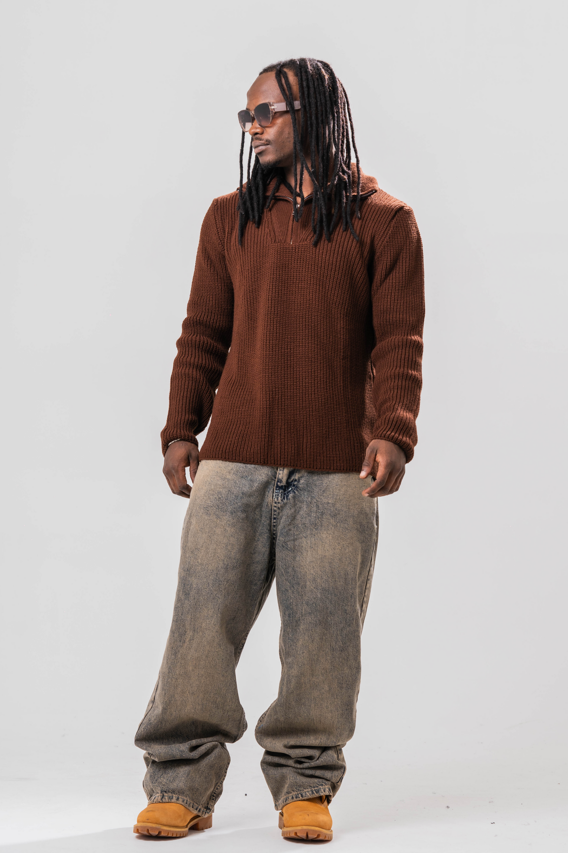 Male model wearing the Sonder Brown Knit Quarter Zip with baggy jeans and sun glasses. In front of a light grey background.