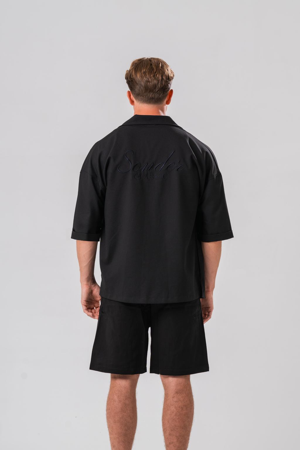 Back view of Male model wearing a white under layer with the Sonder Classic Black oversized shirt and shorts, in front of a light grey background
