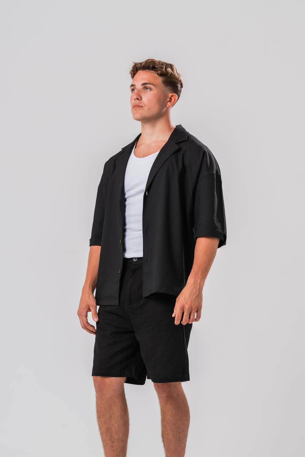 Side view of Male model wearing a white under layer with the Sonder Classic Black oversized shirt and shorts, in front of a light grey background