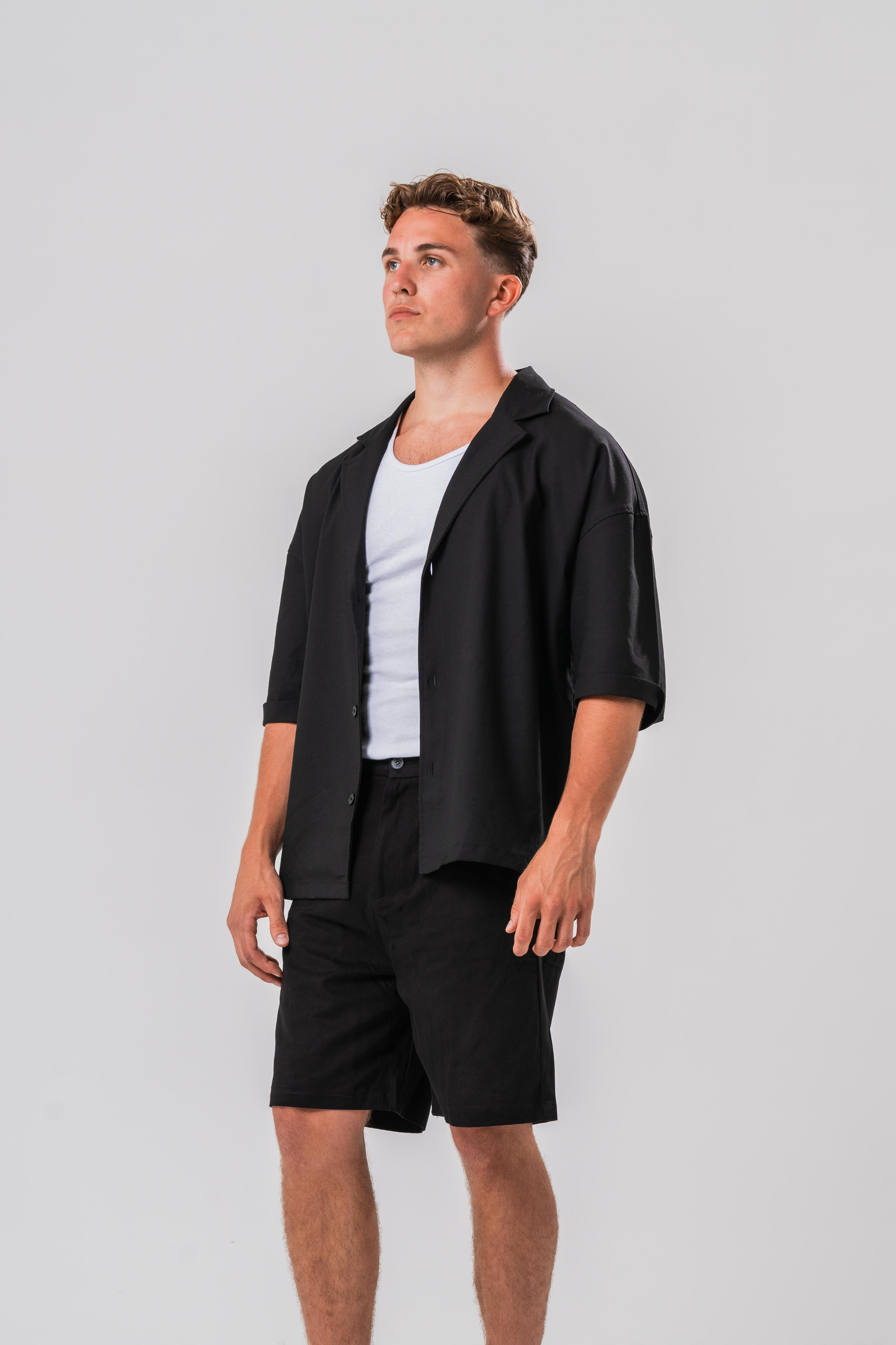 Side view of Male model wearing a white under layer with the Sonder Classic Black oversized shirt and shorts, in front of a light grey background