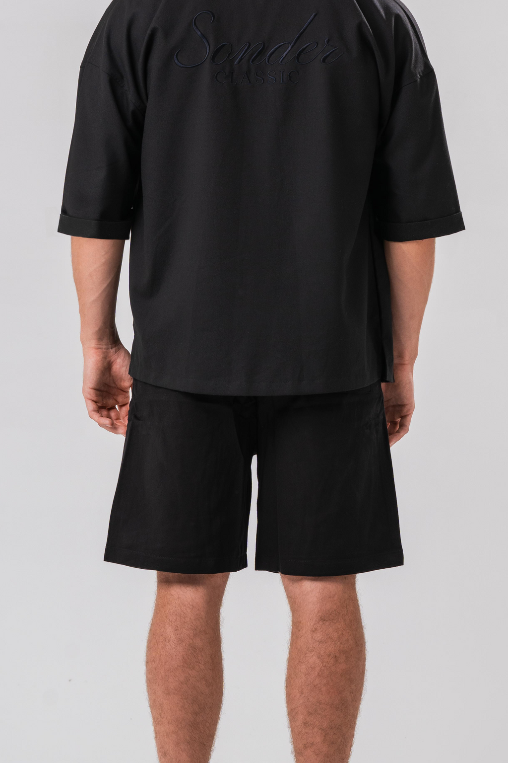 Back view of Male model wearing the Sonder Classic Black oversized shirt and shorts, in front of a light grey background