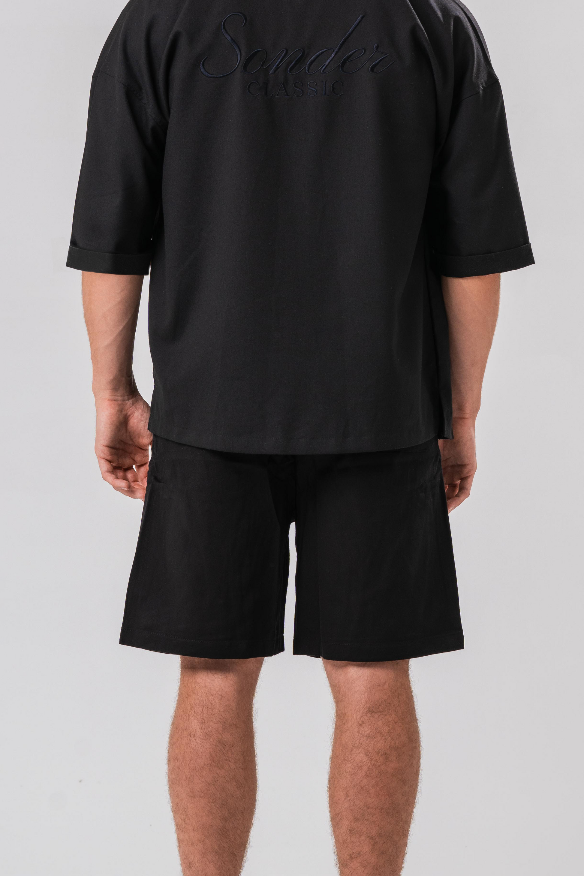 Back view of Male model wearing the Sonder Classic Black oversized shirt and shorts, in front of a light grey background