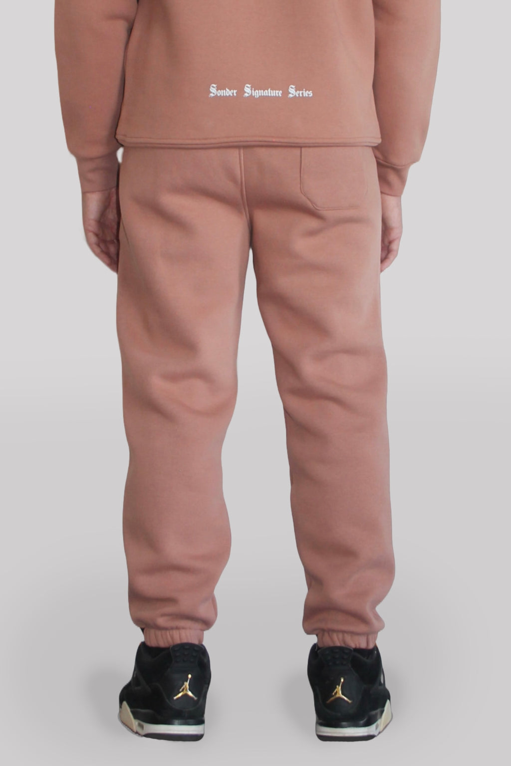 Back view of Male model wearing the Sonder Copper Signature Series Sweatpants paired with the Copper Signature Series Sweatshirt. In front of a light grey background