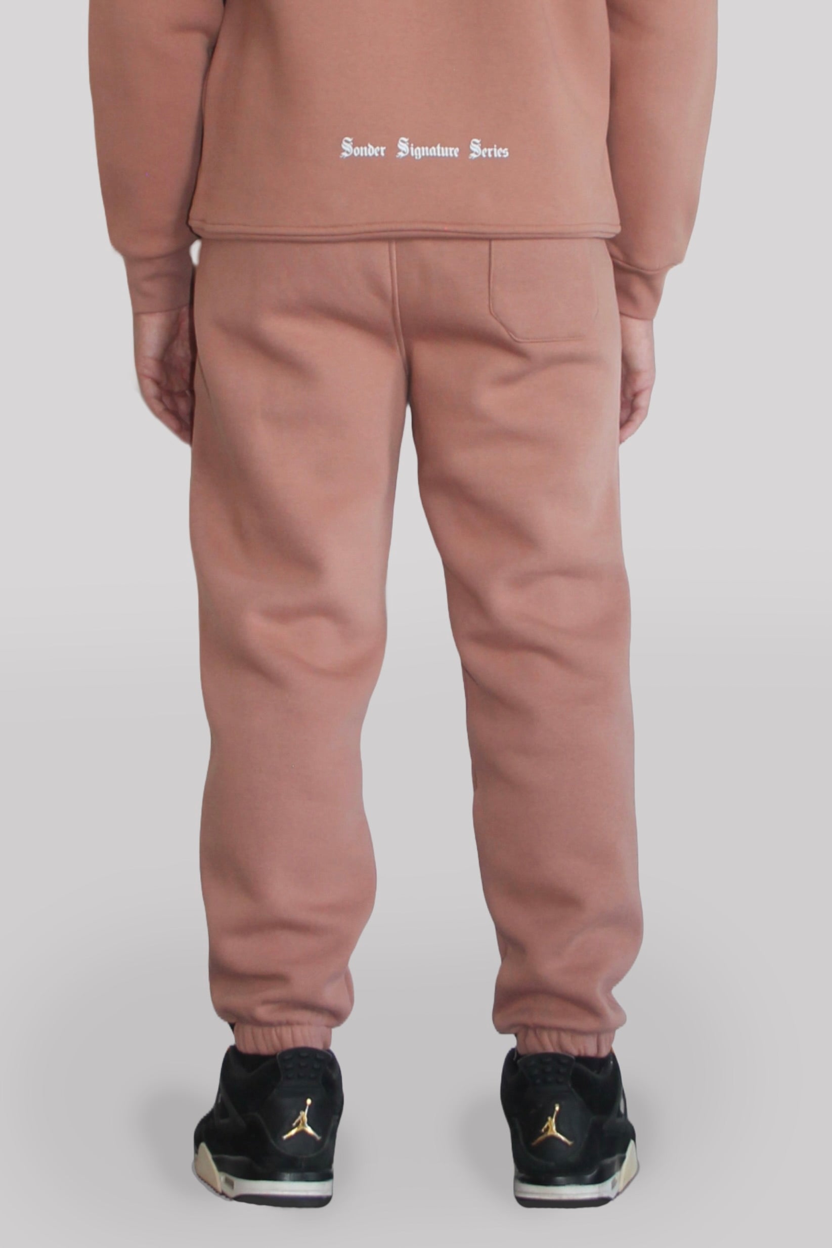 Back view of Male model wearing the Sonder Copper Signature Series Sweatpants paired with the Copper Signature Series Sweatshirt. In front of a light grey background