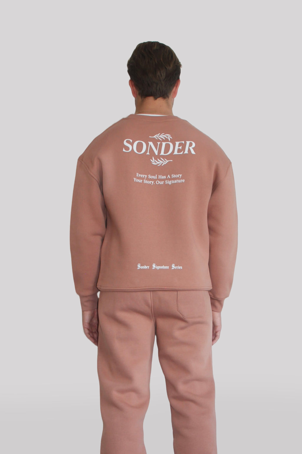 Back view of Male model wearing the Sonder Copper Signature Series Sweatshirt with SONDER branding paired with the Copper Signature Series Sweatpants. In front of a light grey background