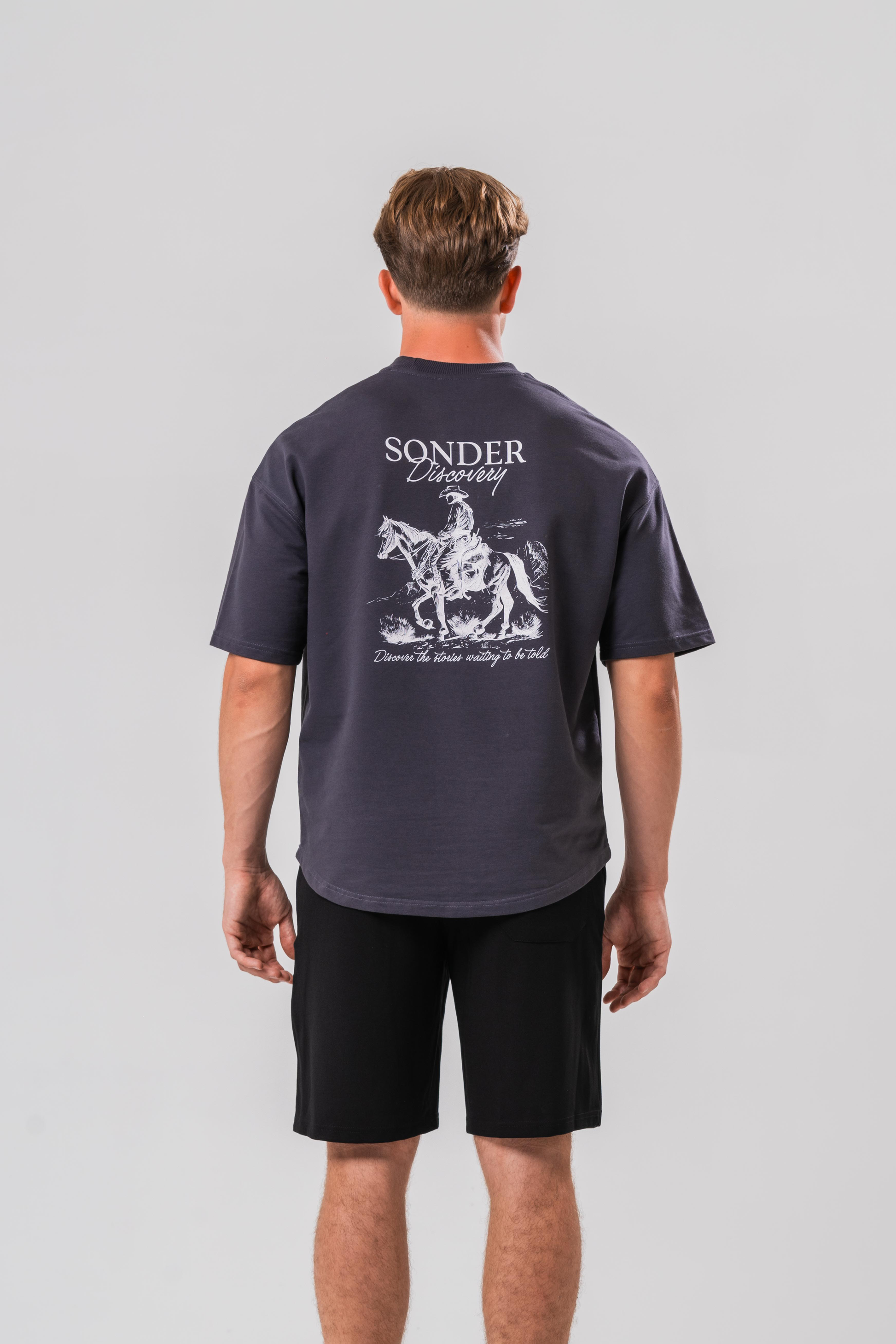 Back of Male model wearing the Sonder Grey cowboy T-shirt and black shorts, in front of a light grey background