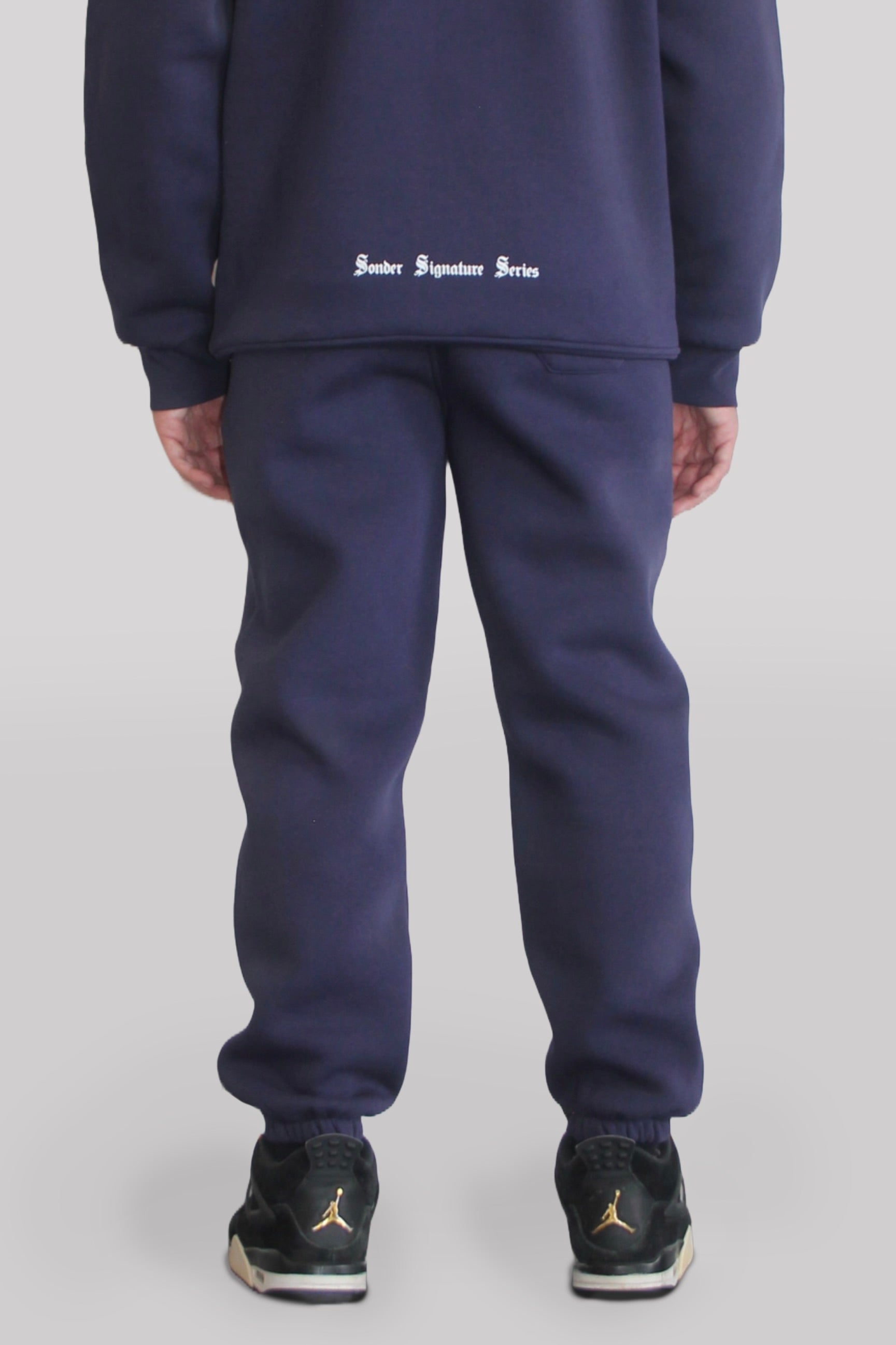 Back view of Male model wearing the Sonder Navy Signature Series Sweatpants paired with the Navy Signature Series Sweatshirt. In front of a light grey background.