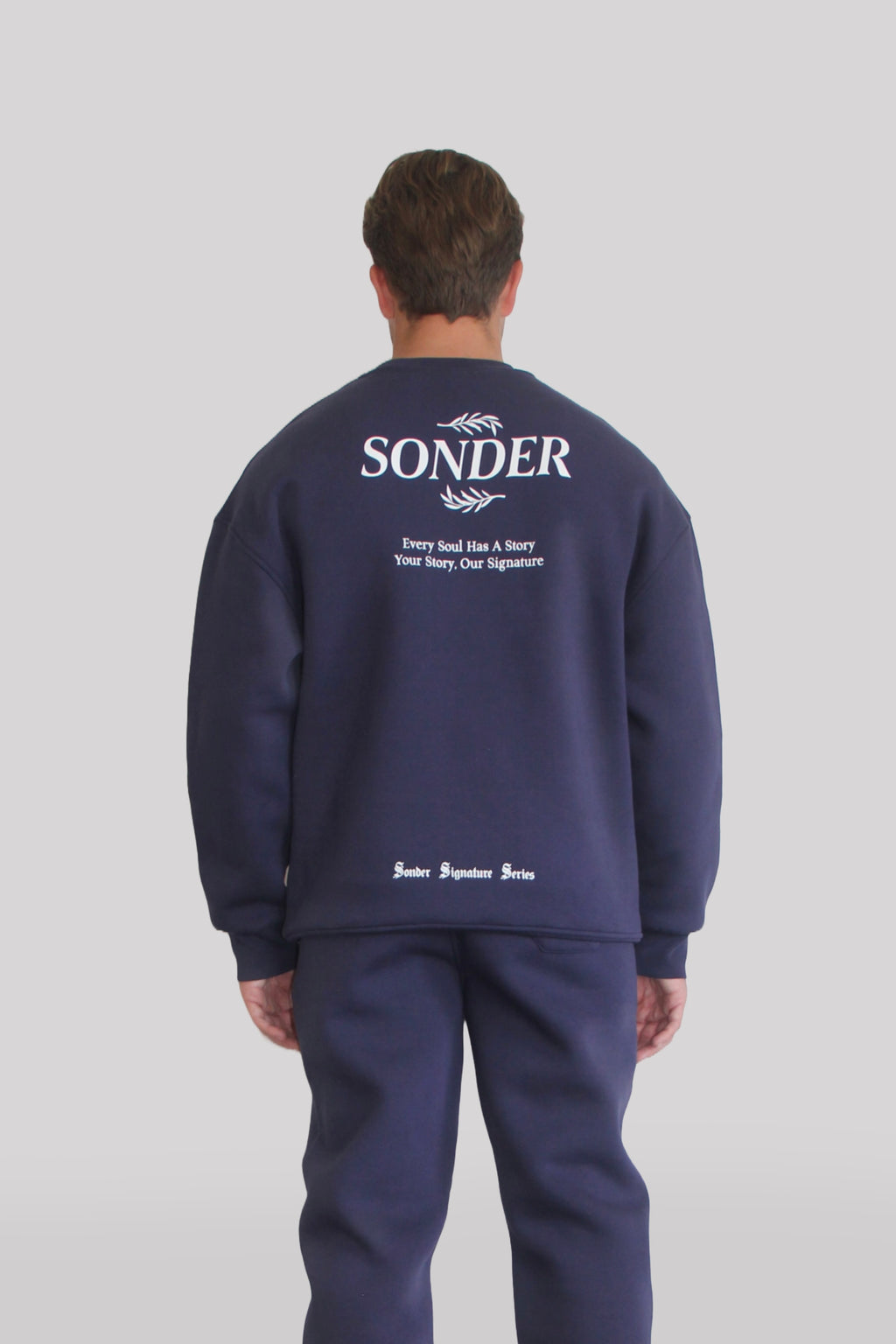 Back view of Male model wearing the Sonder Navy Signature Series Sweatshirt with SONDER branding paired with the Navy Signature Series Sweatpants. In front of a light grey background