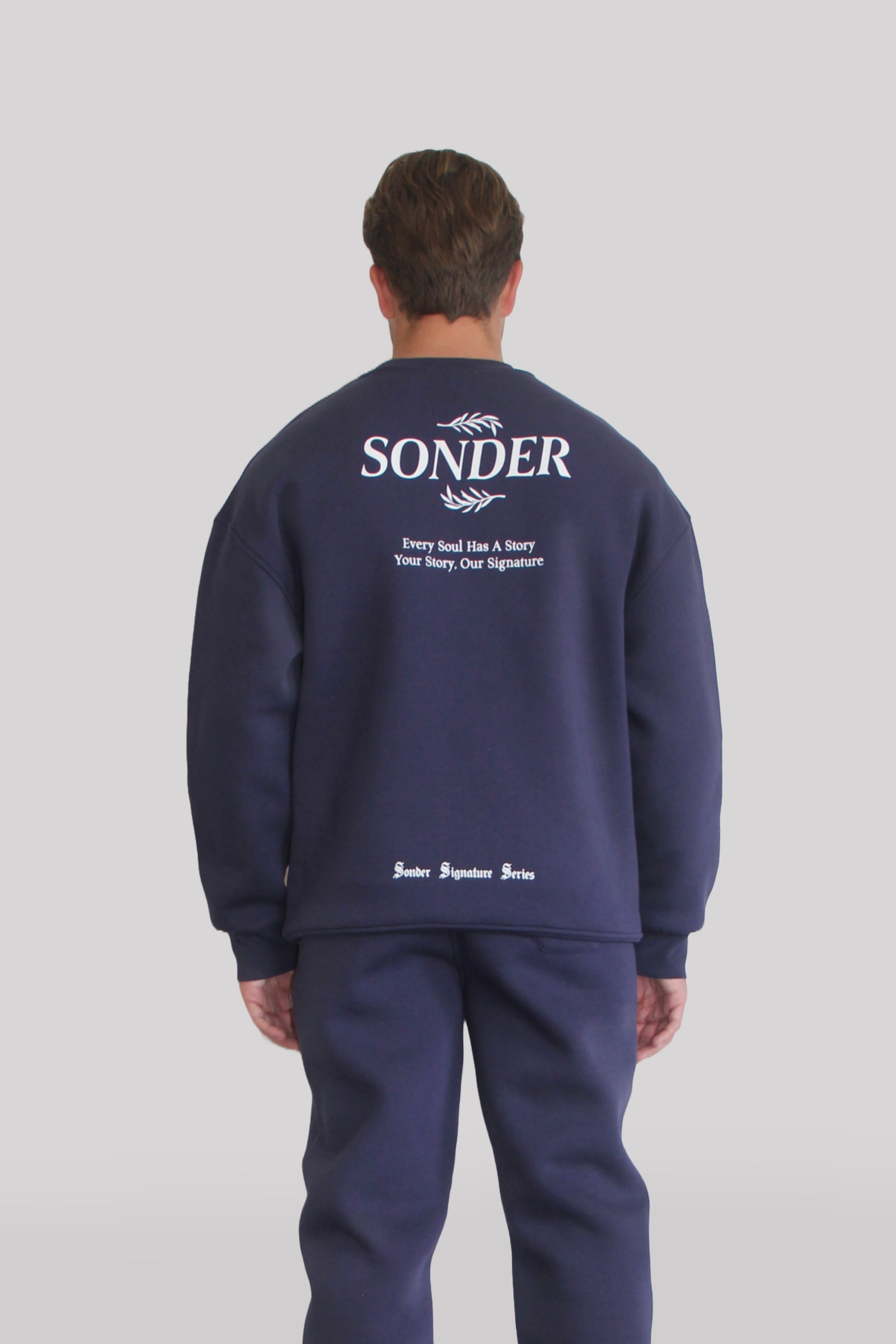 Back view of Male model wearing the Sonder Navy Signature Series Sweatshirt with SONDER branding paired with the Navy Signature Series Sweatpants. In front of a light grey background