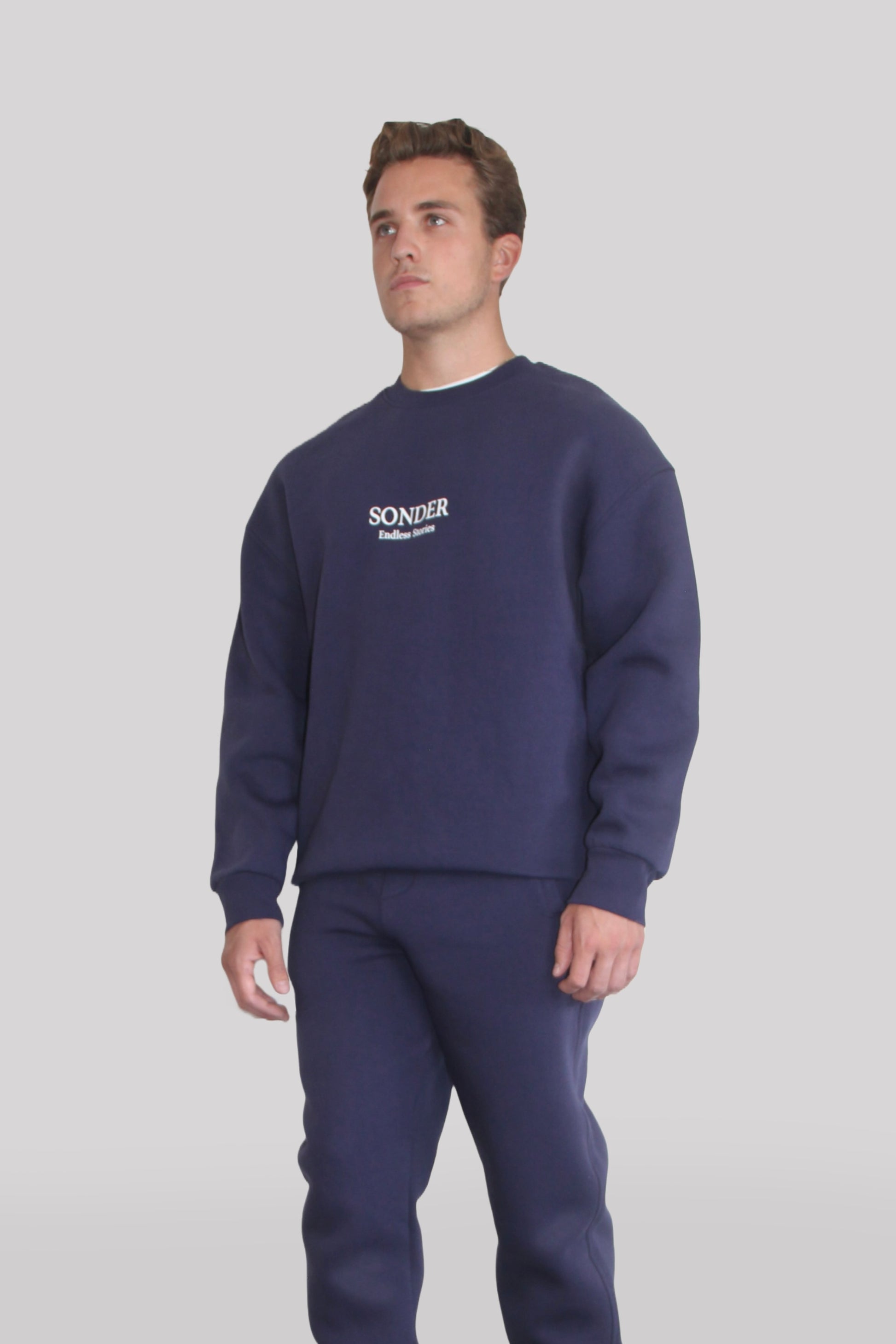 Side view of Male model wearing the Sonder Navy Signature Series Sweatshirt with SONDER branding paired with the Navy Signature Series Sweatpants. In front of a light grey background.