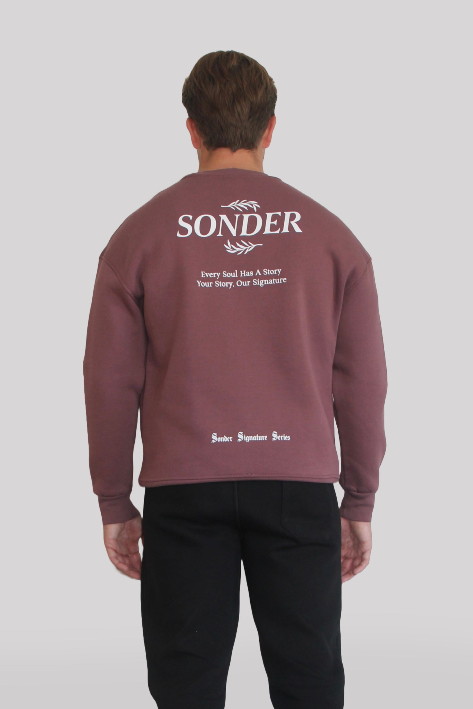 Back view of Male model wearing the Sonder Plum Signature Series Sweatshirt with SONDER branding paired with the Black Signature Series Sweatpants. In front of a light grey background