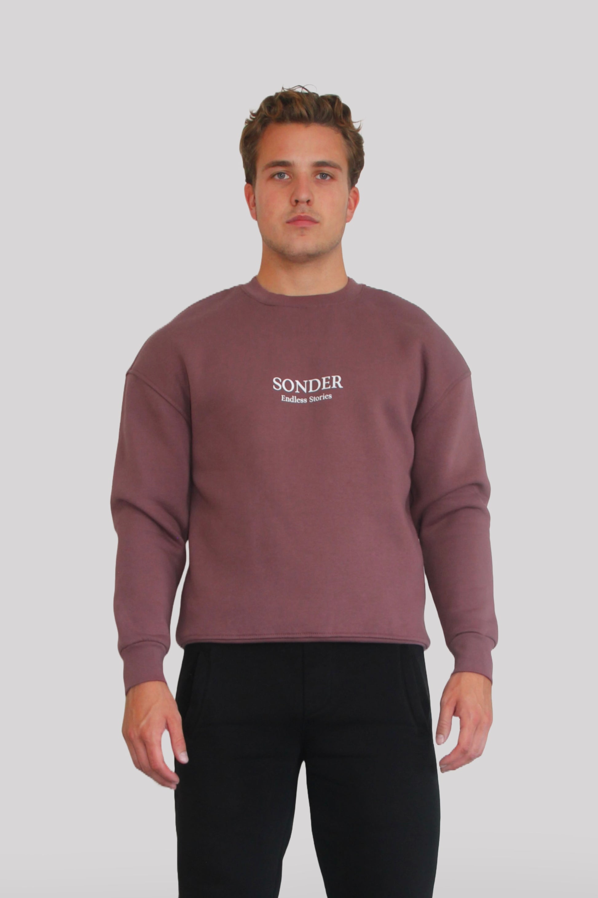 Male model wearing the Sonder Plum Signature Series Sweatshirt with SONDER branding paired with the Black Signature Series Sweatpants. In front of a light grey background