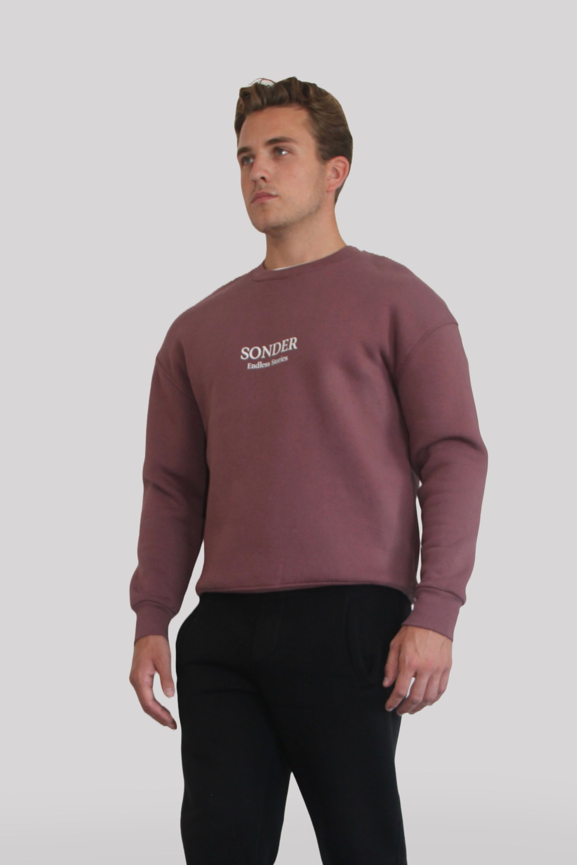 Side view of Male model wearing the Sonder Plum Signature Series Sweatshirt with SONDER branding paired with the Black Signature Series Sweatpants. In front of a light grey background