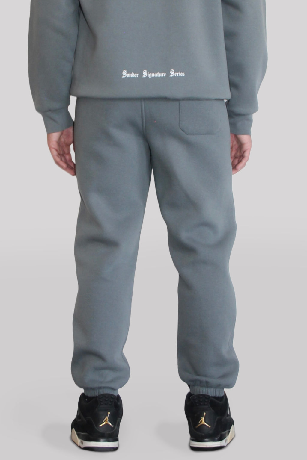 Back view of Male model wearing the Sonder Shadow Grey Signature Series Sweatpants paired with the Shadow Grey Signature Series Sweatshirt. In front of a light grey background.
