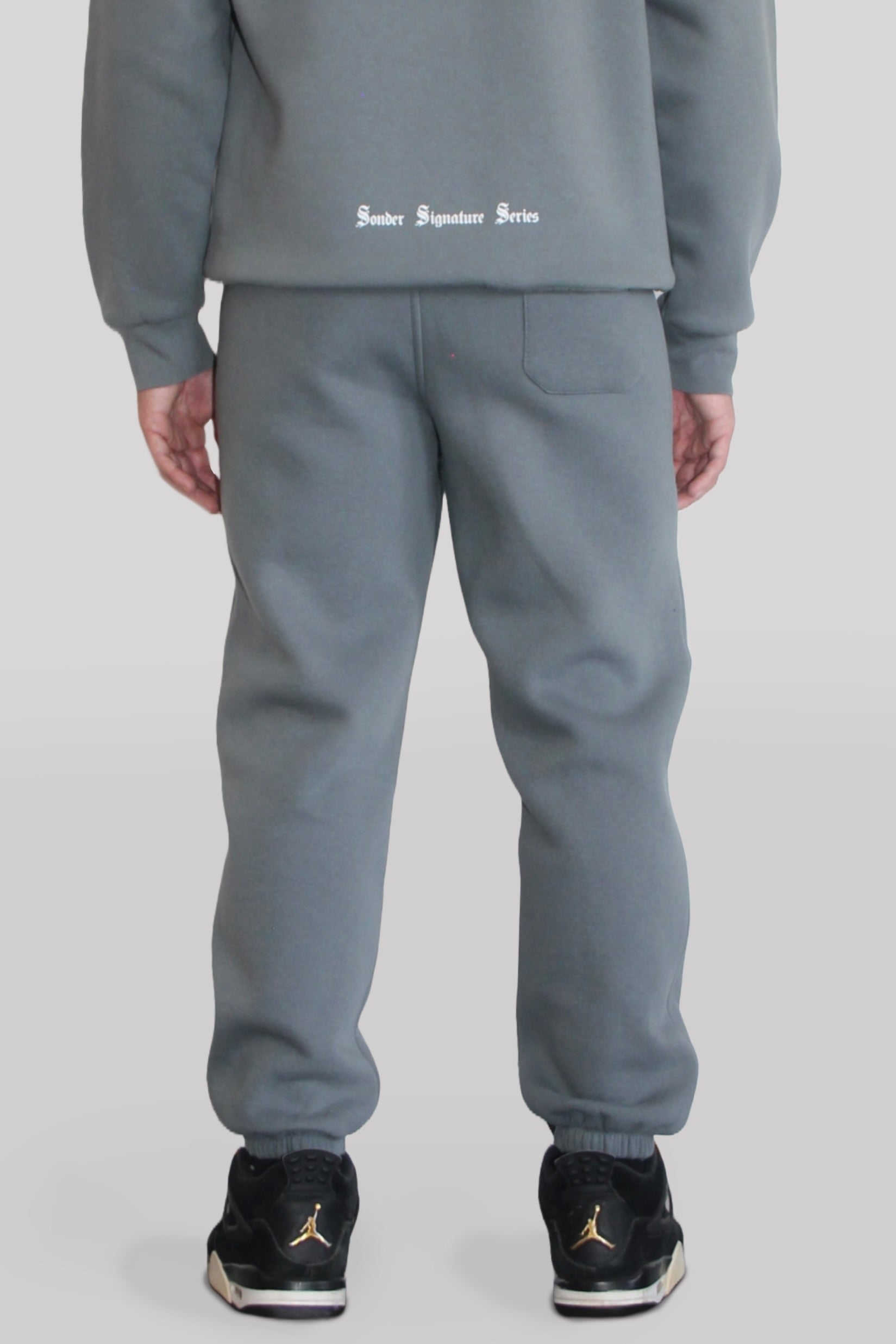 Back view of Male model wearing the Sonder Shadow Grey Signature Series Sweatpants paired with the Shadow Grey Signature Series Sweatshirt. In front of a light grey background.
