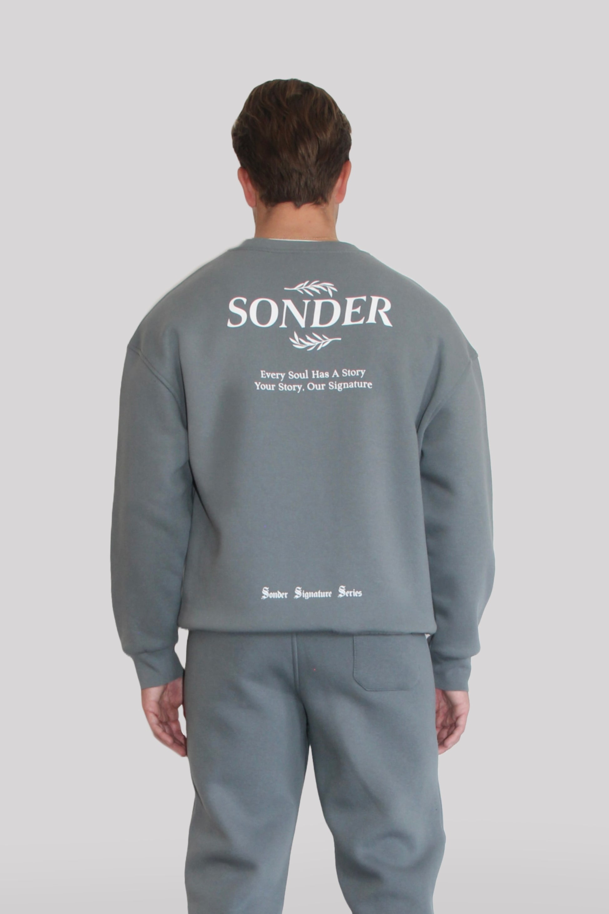 Back view of Male model wearing the Sonder Shadow Grey Signature Series Sweatshirt with SONDER branding paired with the Shadow Grey Signature Series Sweatpants. In front of a light grey background