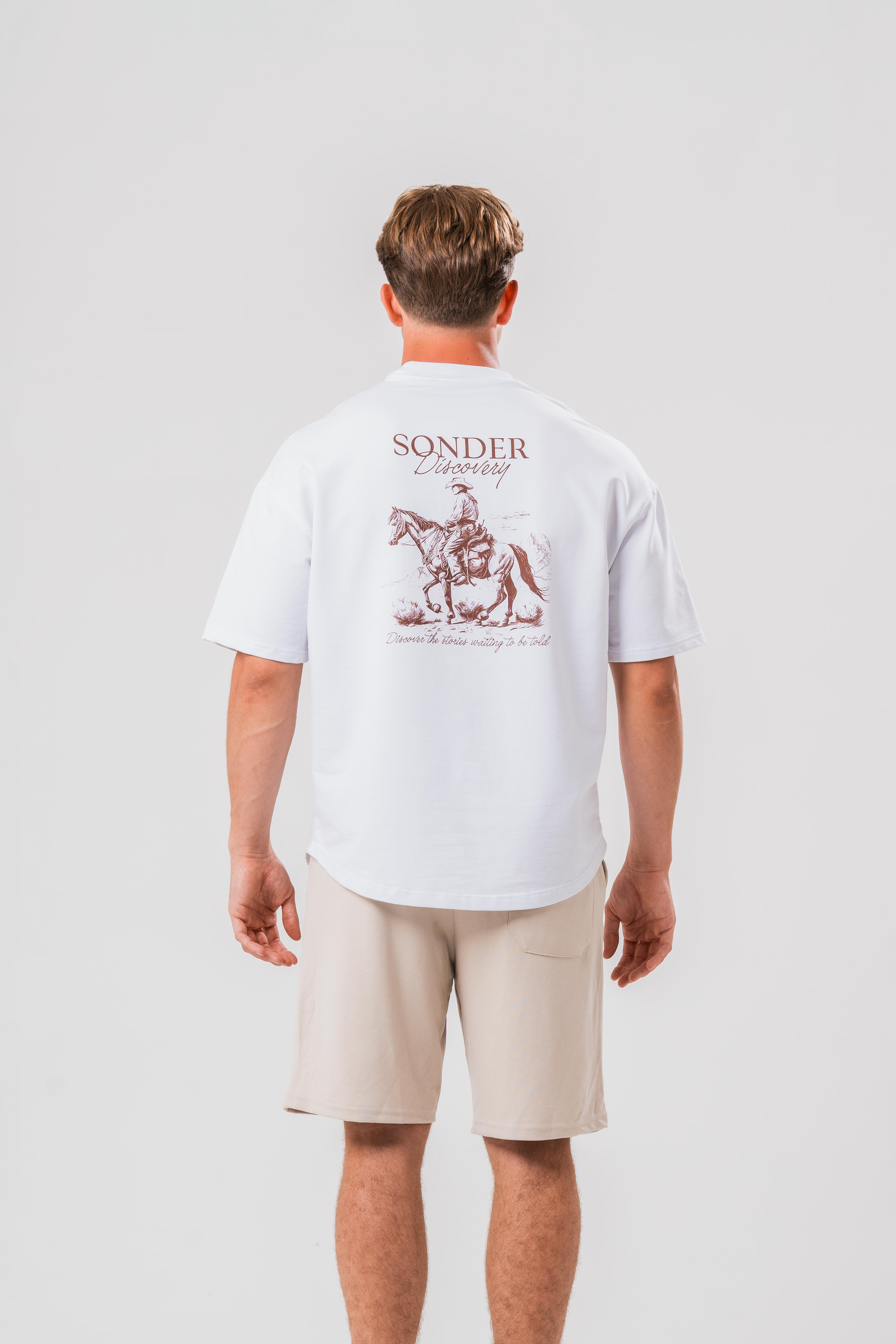 Back of Male model wearing the Sonder White cowboy T-shirt and beige shorts, in front of a light grey background