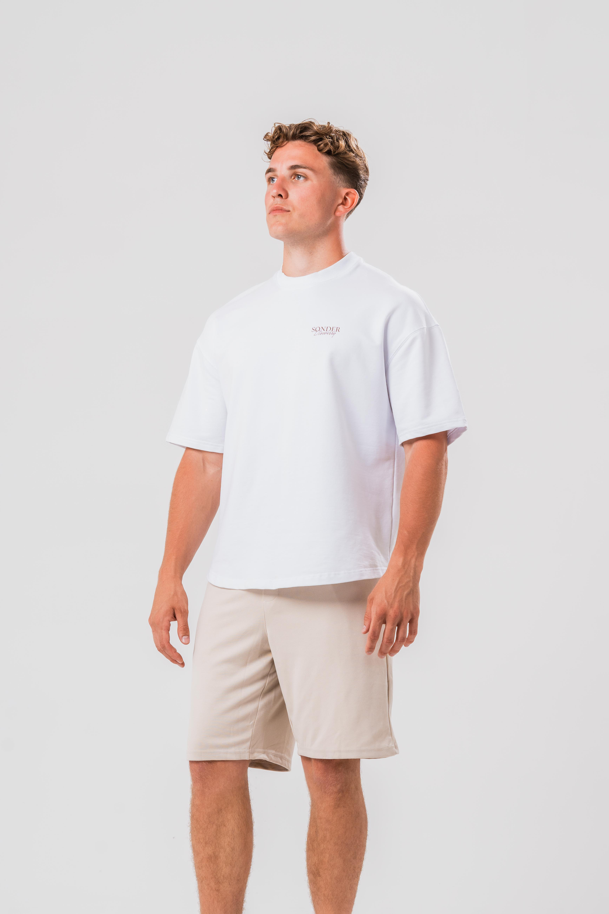 Side view of Male model wearing the Sonder White cowboy T-shirt and beige shorts, in front of a light grey background