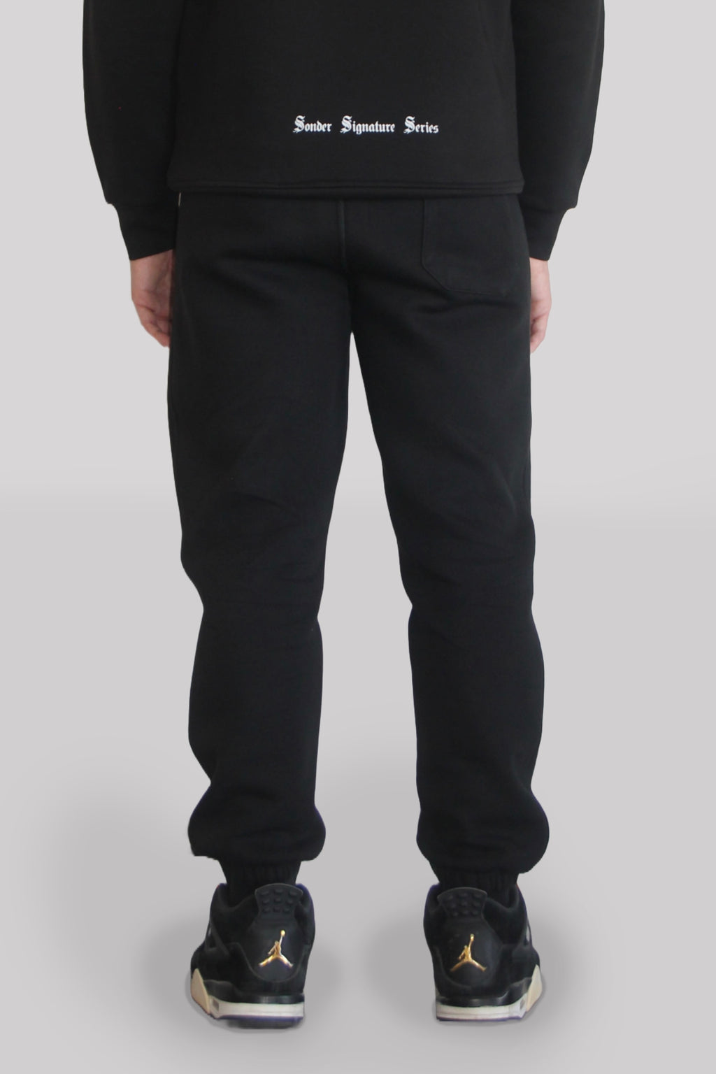 Back view of Male model wearing the Sonder black Signature Series Sweatpants paired with the Black Signature Series Sweatshirt. In front of a light grey background
