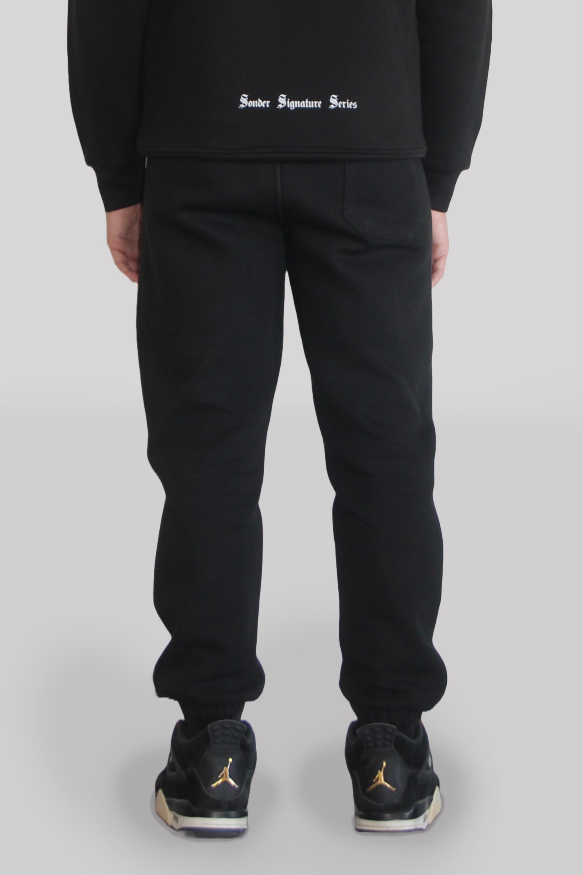 Back view of Male model wearing the Sonder black Signature Series Sweatpants paired with the Black Signature Series Sweatshirt. In front of a light grey background